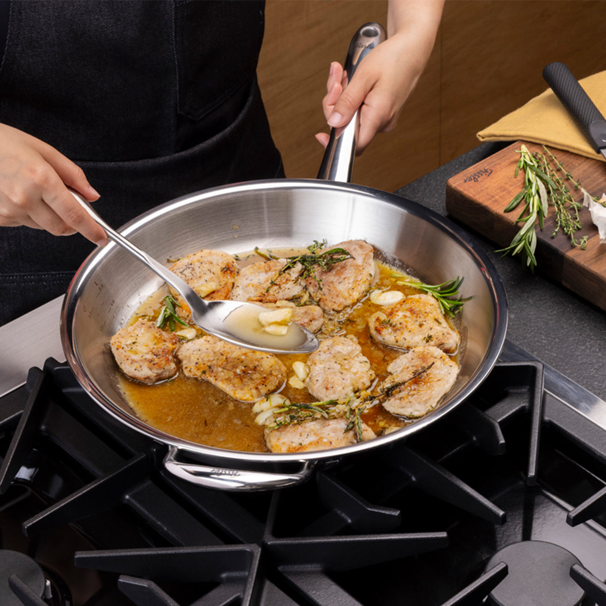 Eine Person bereitet in der Fissler M5 Pro-Ply®-Pfanne aus recyceltem Edelstahl auf einem Gasherd perfekt gewürzte Fleischmedaillons mit frischen Kräutern und Knoblauch zu. Mit einem Löffel übergießt der Koch das Fleisch gekonnt mit einer reichhaltigen Soße. Die Fissler M5 Pro-Ply®-Pfanne spiegelt unsere Tradition der Präzisionstechnik und des zeitlosen deutschen Designs wider – induktionsgeeignet für den Einsatz auf allen Kochfeldern und mühelose Reinigung für den täglichen Gebrauch. Im Hintergrund sind