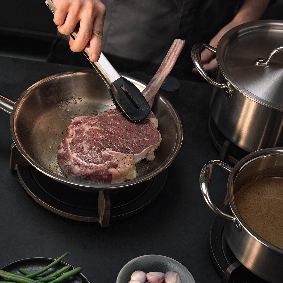 Ein Koch brät gekonnt ein großzügiges Steak in der Fissler M5 Pro-Ply®-Pfanne an, die aus recyceltem Edelstahl gefertigt ist und sich durch langlebige Qualität sowie hervorragende Wärmeverteilung auszeichnet. Die Szene unterstreicht die renommierte deutsche Handwerkskunst von Fissler: Neben zwei glänzenden Fissler-Töpfen auf dem Herd kommen eine Zange und ein Pfannenwender zum Einsatz. Frische grüne Bohnen und Knoblauchzehen liegen auf einer eleganten, dunklen Arbeitsplatte und unterstreichen das Konzept