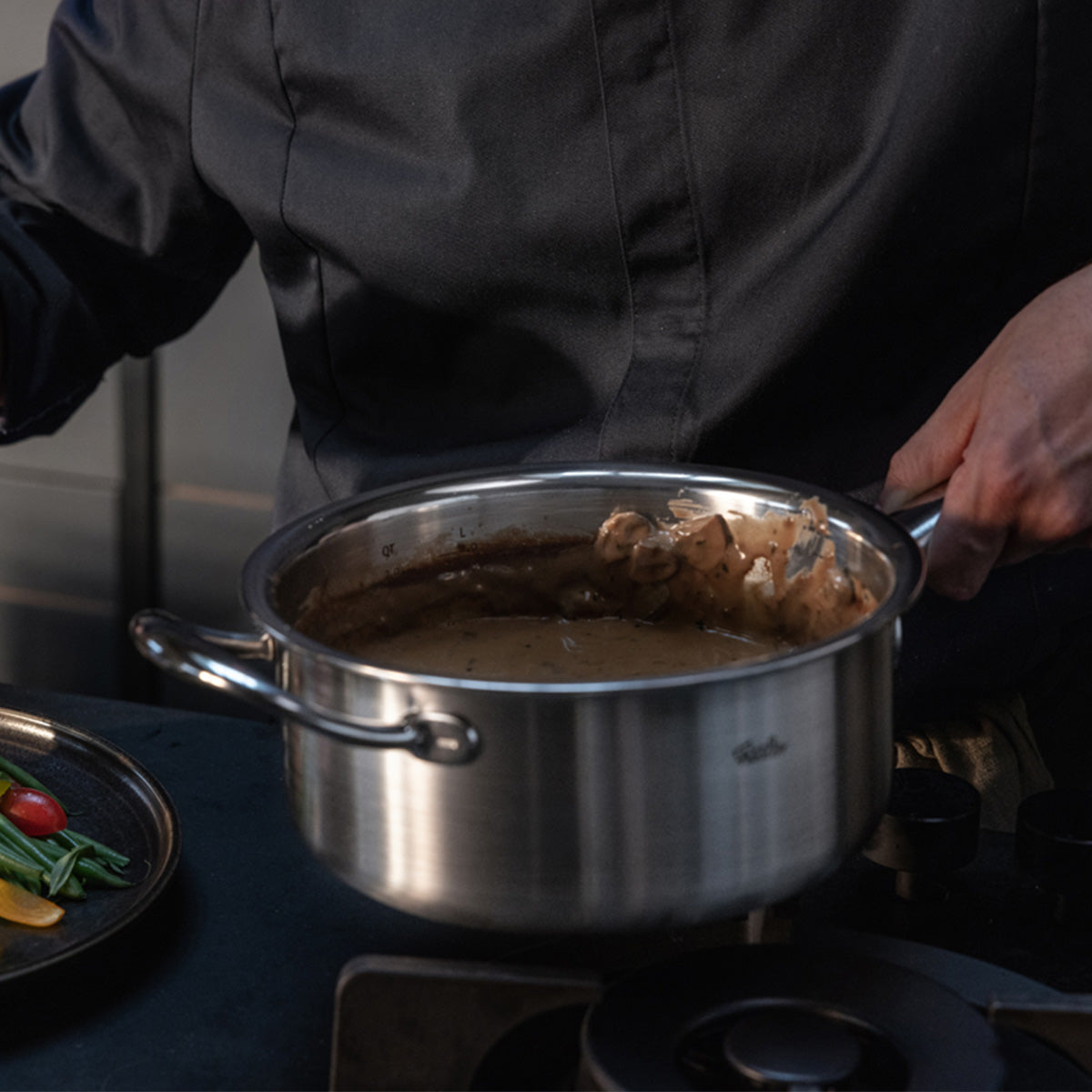 Eine Person in einer dunklen Kochjacke rührt gekonnt mit einem Pfannenwender eine cremige braune Soße in einem Fissler M5 Pro-Ply®-Edelstahltopf mit Metalldeckel aus dem 3-teiligen Set, der auf dem Herd steht. Das hochwertige Kochgeschirr, hergestellt in Deutschland und bekannt für seine Präzisionstechnik und sein zeitloses Design, eignet sich sowohl für Induktionskochfelder als auch für Backöfen. Daneben unterstreicht ein Teller mit frischem Gemüse die Vielseitigkeit und Qualität, die die traditionelle
