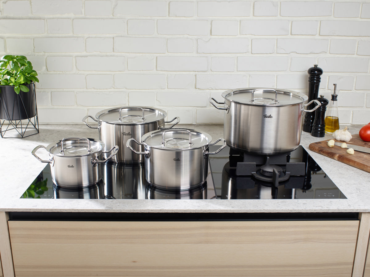 Fissler Original-Profi Collection® 5-tlg. Set auf modernem Herd, umgeben von Gemüse. Edelstahl, "Made in Germany", spülmaschinenfest.