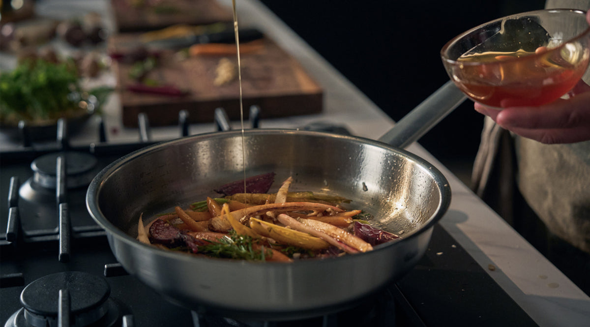 Eine Hand gießt behutsam Flüssigkeit aus einer Glasschüssel in eine hochwertige Fissler-Edelstahlpfanne auf dem Herd, in der frisches, gleichmäßig geschnittenes Gemüse gekonnt angebraten wird. Die Szene verdeutlicht die bekannte deutsche Präzision von Fissler und das zeitlose Design der Kochgeschirrserie.