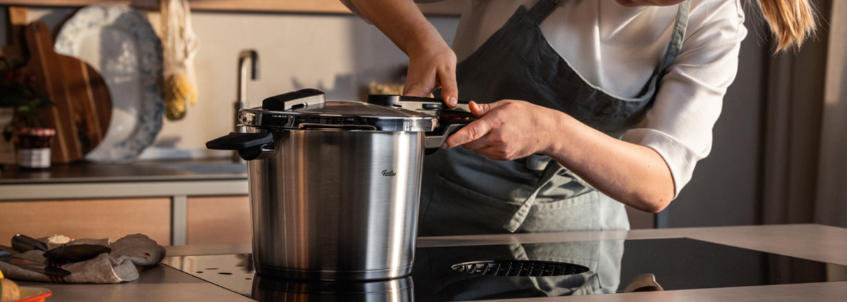 In einer eleganten, modernen Küche verschließt eine Person, die eine Schürze trägt, den Deckel eines Fissler-Edelstahl-Schnellkochtopfs und unterstreicht damit die berühmte deutsche Ingenieurskunst und das zeitlose Design der Marke.