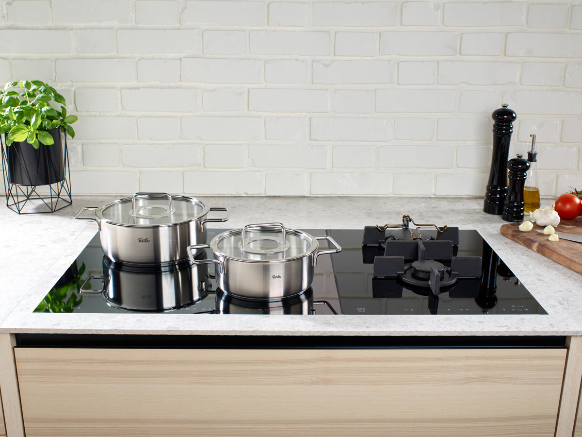 A modern kitchen counter with two Fissler Pure Collection pots, fresh herbs, and vegetables on an elegant black glass stovetop.