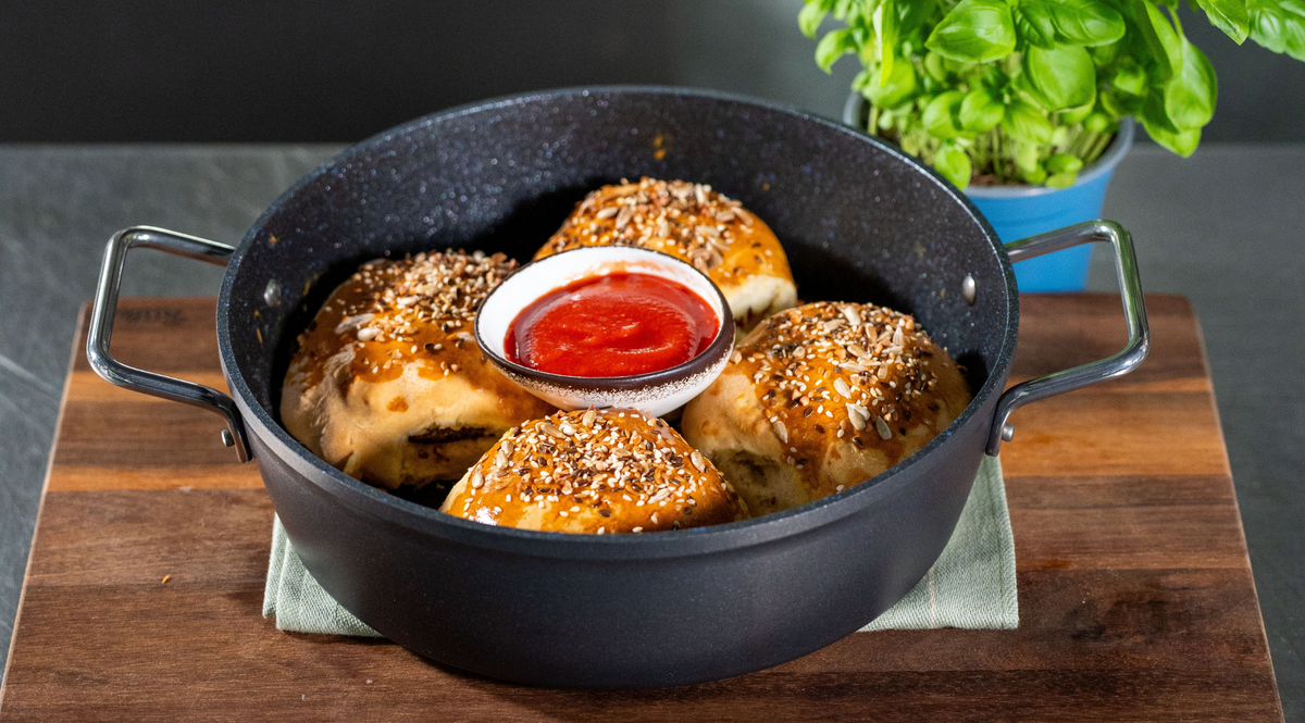 Vier mit Sesam bestreute Brötchen sind kunstvoll in einem schwarzen Fissler-Topf angeordnet, in dessen Mitte eine Schale mit leuchtend roter Dip-Sauce steht – alles elegant auf einem Holzbrett präsentiert. Eine kleine Basilikumpflanze im Topf sorgt im Hintergrund für einen frischen Akzent und unterstreicht Fisslers Engagement für Qualität und zeitloses Design in Ihrer Küche.