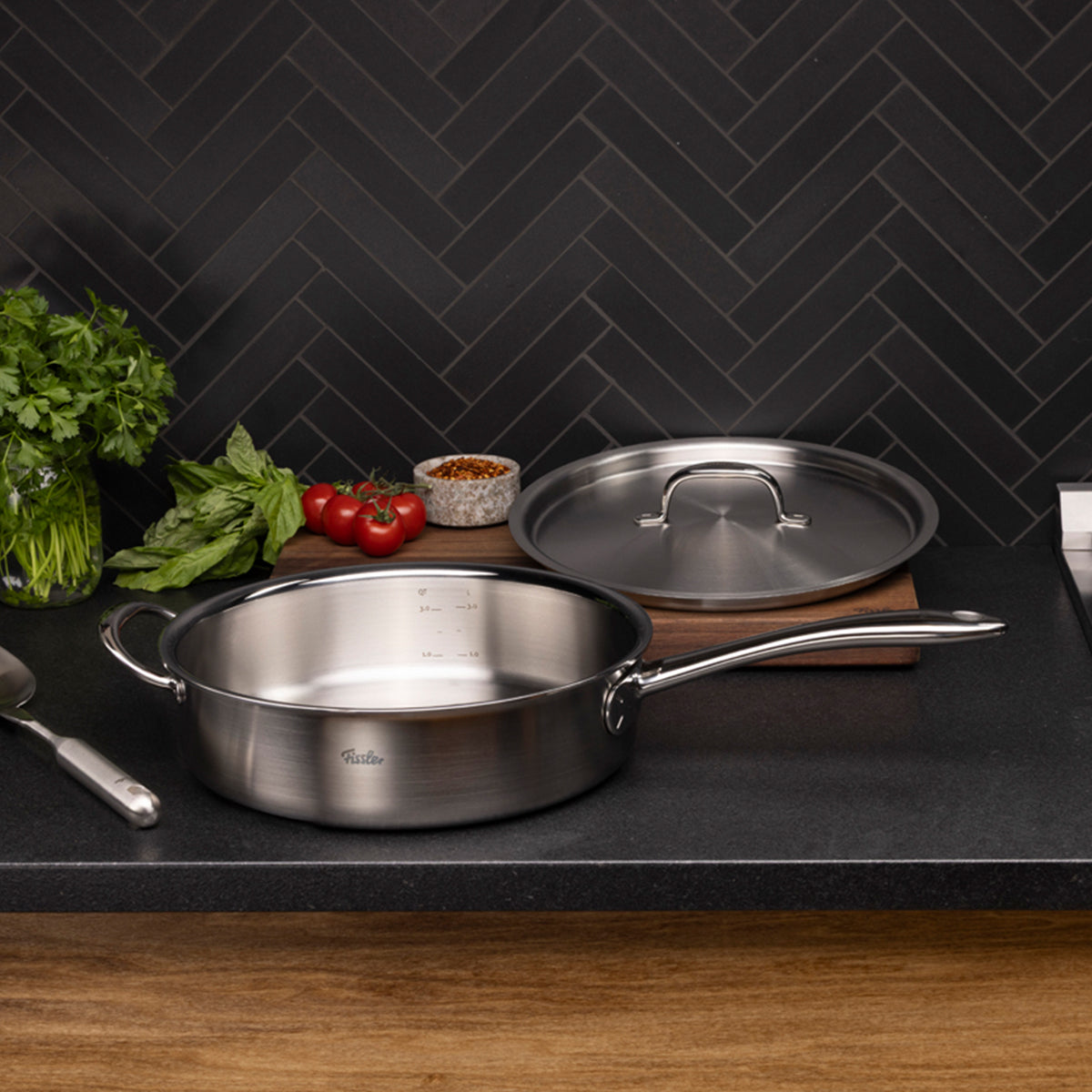 Die Fissler M5 Pro-Ply® Sautépfanne mit Metalldeckel aus recyceltem Edelstahl steht auf einer eleganten, dunklen Küchenarbeitsplatte. Im Vordergrund liegen frische Kräuter, Tomaten und kleine Schälchen mit Gewürzen vor einer stilvollen schwarzen Fliesenwand im Fischgrätenmuster. Die hochwertige Sautépfanne von Fissler, gegründet 1845 und bekannt für langlebige Präzision „Made in Germany“, ist induktionsgeeignet, spülmaschinenfest und verbindet innovative Nachhaltigkeit mit zeitlosem Design – ein Beispiel