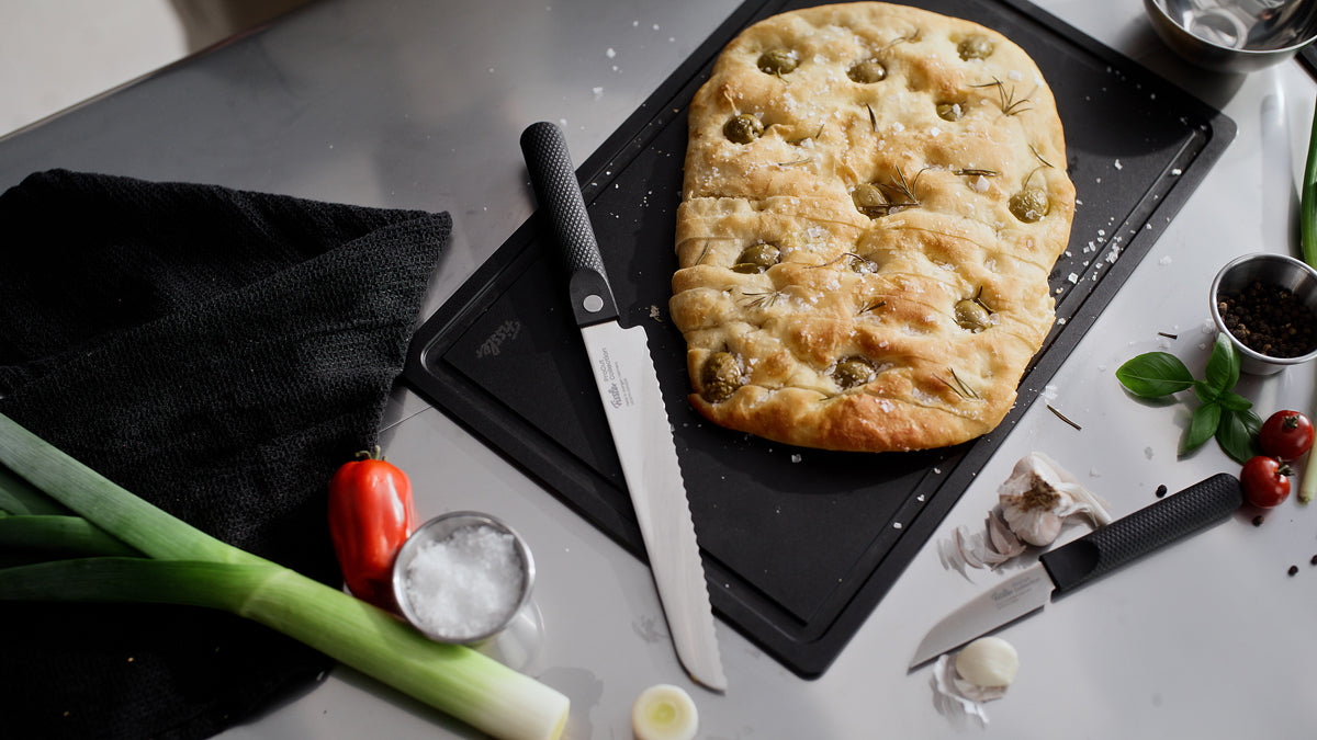 A rectangular olive focaccia sits on a black cutting board, accompanied by the Fissler ProCut Brotmesser 21 cm—crafted with extraharter Stahl and precision Wellenschliffklinge for effortless slicing—with a matching paring knife nearby. The arrangement includes fresh leeks, tomatoes, garlic cloves, salt, basil leaves, peppercorns in a metal cup, and a black cloth. This scene showcases Fissler’s commitment to German-engineered quality and timeless kitchen design since 1845.