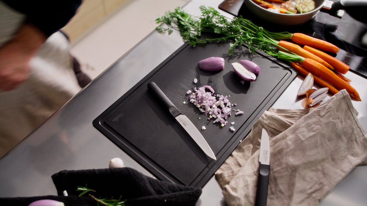 On a kitchen counter, the Fissler ProCut Universalmesser 14 cm and a small Fissler kitchen knife, both crafted from extra-hardened steel and designed for exceptional precision and durability, are displayed alongside a cutting board with finely chopped shallots, whole and halved shallots, fresh carrots, and green herbs. A person stands nearby, partially out of frame—showcasing the timeless quality and innovative engineering that define Fissler’s German-made cookware.