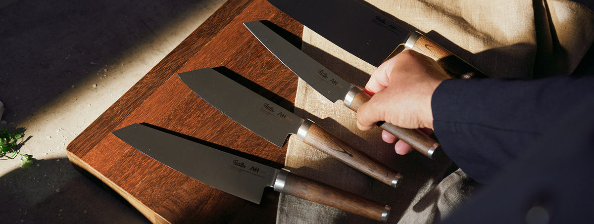 Close-up of a hand selecting a Fissler chef's knife with a wooden handle from four knives lying on a wooden board, with the sun highlighting the craftsmanship.