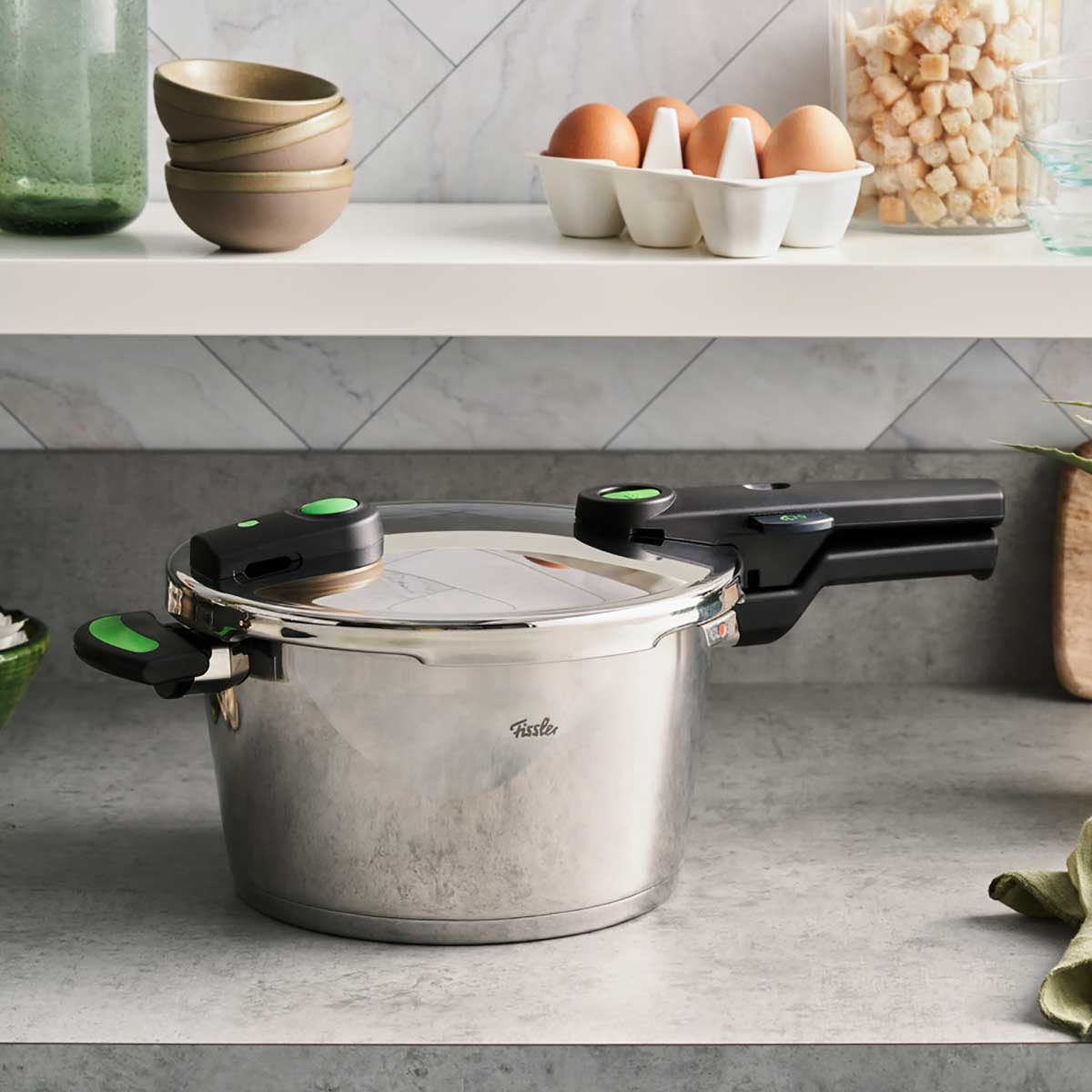 Der Fissler Vitaquick® Green Schnellkochtopf mit markanten Griffen auf grauer Platte, umgeben von Küchenaccessoires.
