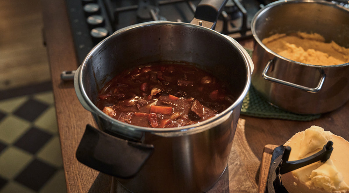 Auf einer Holzarbeitsplatte neben dem Herd steht ein Fissler-Edelstahltopf, der mit präziser Technik und zeitlosem Design gefertigt wurde und einen herzhaften Rindfleischeintopf mit großzügigen Fleisch- und Gemüsestücken enthält. Daneben steht ein weiterer Fissler-Topf mit cremig-gelbem Kartoffelpüree, dazu ein Teller mit Butter – ein Beispiel für die Langlebigkeit und Innovationskraft der in Deutschland hergestellten Fissler-Kochgeschirre.