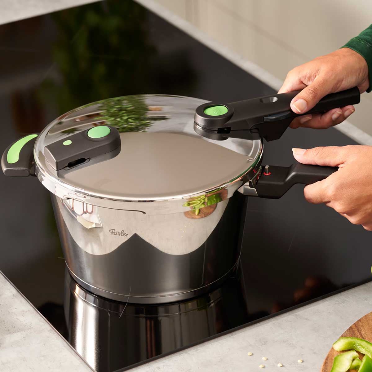 Eine Person befestigt den Deckel eines Fissler Vitaquick® Green Schnellkochtopfes mit schwarz-grünen Griffen auf einem Herd in einer eleganten, modernen Küche.