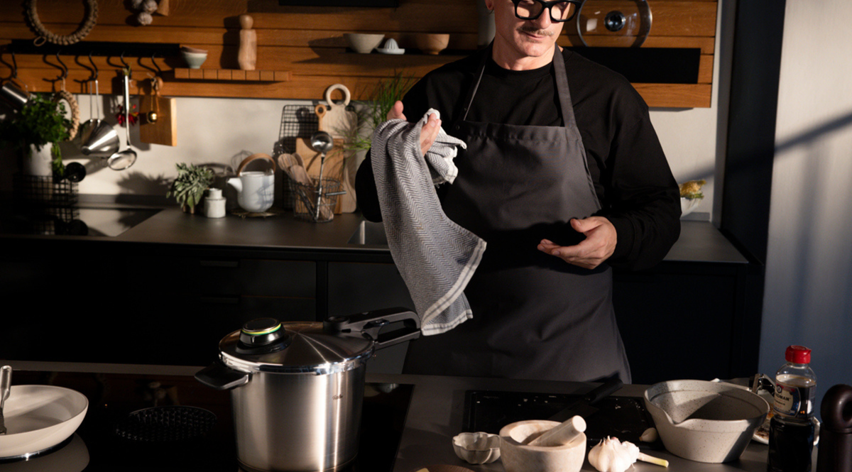 Eine Person in einer dunklen Küche steht neben einem Fissler Schnellkochtopf auf dem Herd, umgeben von hochwertigem Kochgeschirr und frischen Zutaten.