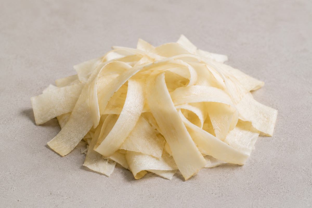 Breite, blasse Streifen aus rohem Yuca oder Maniok sind kunstvoll auf einem hellen Untergrund angeordnet und verdeutlichen, wie präzise sich mit den fachmännisch entwickelten Küchenwerkzeugen von Fissler schneiden lässt – perfekt für alle, die bei der Zubereitung von Speisen Wert auf zeitloses Design und langlebige Qualität legen.