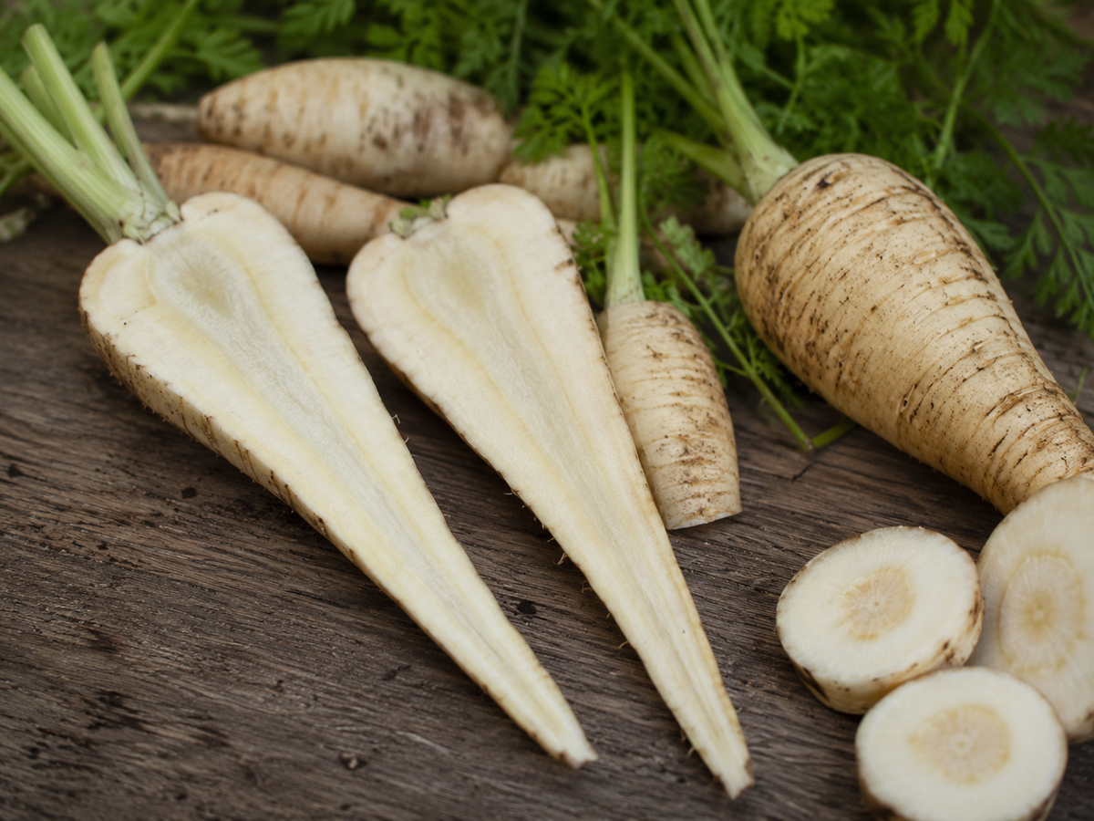 Mehrere frische Pastinaken mit grünem Laub liegen auf einer Holzunterlage und sind wunderschön arrangiert neben präzise geschnittenen runden Scheiben und einer halbierten Pastinake, die ihren inneren Aufbau offenbart – eine einladende Kulisse für die Zubereitung hochwertiger Zutaten mit dem meisterhaft gefertigten Kochgeschirr von Fissler.