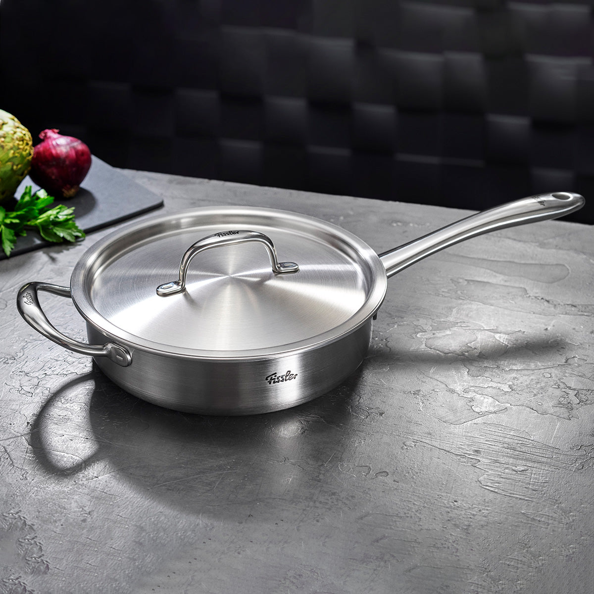 Fissler M5 Pro-Ply Sautépfanne aus recyceltem Edelstahl auf grauer Platte, Gemüse und Artischocke im Hintergrund, induktionstauglich.