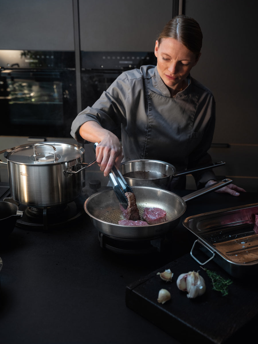 A chef fries a steak in a Frying Pan from the Fissler Original-Profi Collection®, surrounded by our high-quality Cookware, herbs, and garlic.