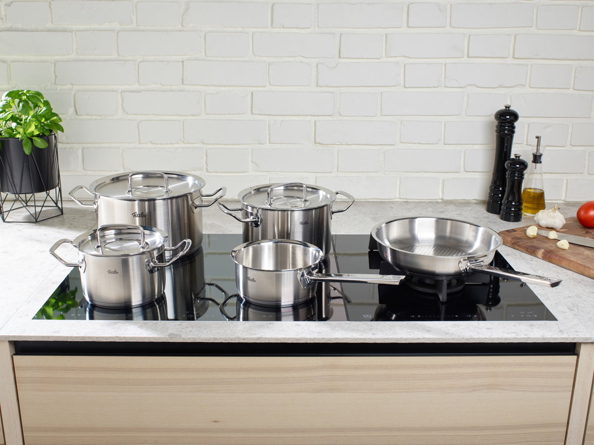 Fissler Original-Profi Collection® 5-teiliges Set: vier rostfreie Töpfe & Pfannen, Premium 18/10 Stahl, auf Induktionsherd, frische Kräuter in der Nähe.