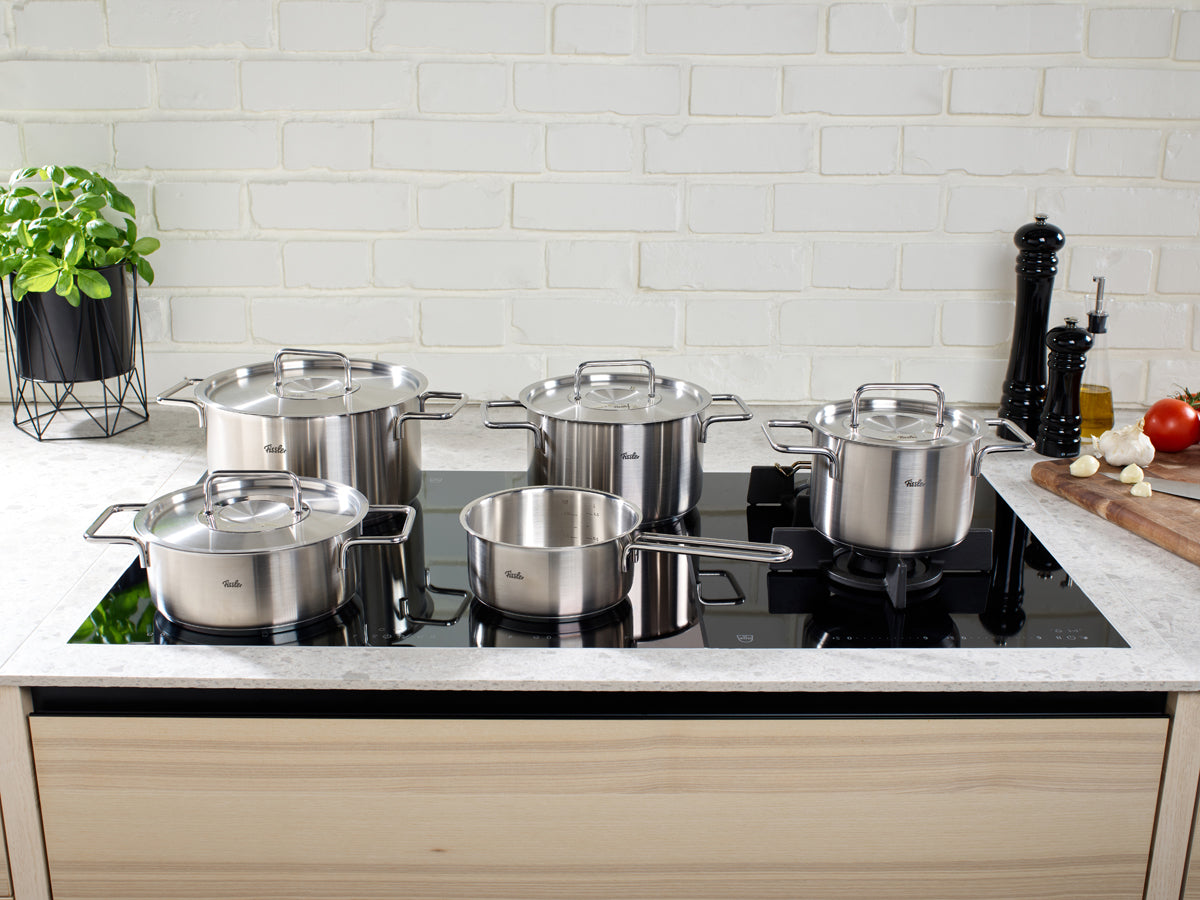 Fissler Pure Collection® 5-tlg. Topf-Set aus Edelstahl auf schwarzem Kochfeld in moderner Küche mit Backsteinwand und Holzschränken.