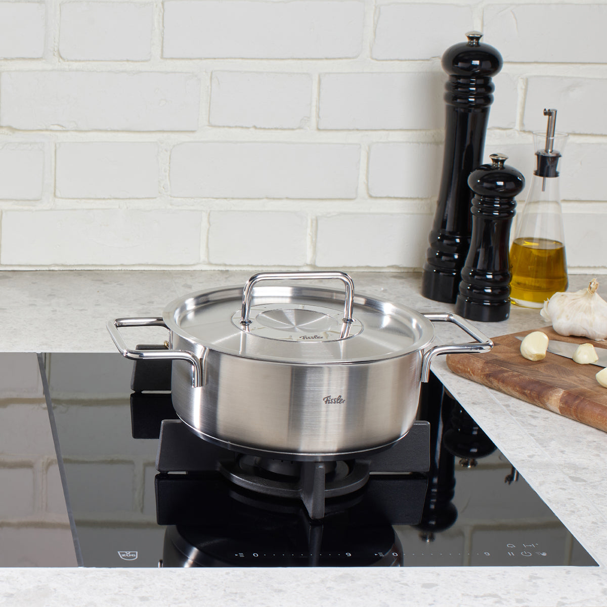 Fissler Pure Collection Bratentopf mit Metalldeckel auf schwarzem Gasherd, umgeben von Küchenutensilien und Knoblauch.