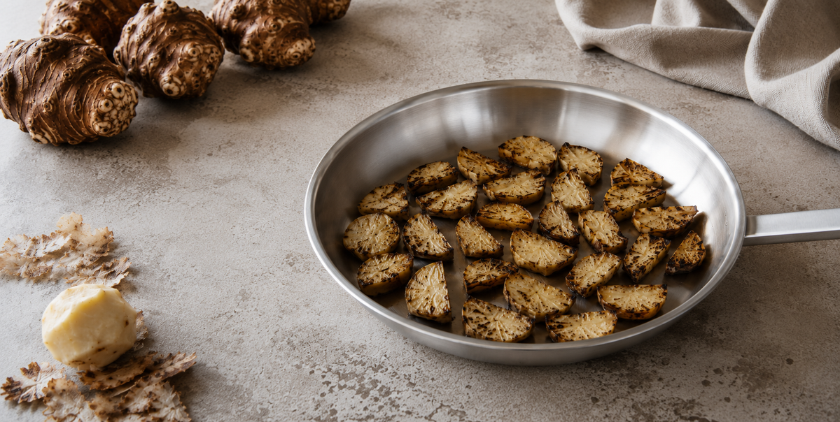 In Scheiben geschnittenes, geröstetes Wurzelgemüse, präsentiert in einer Fissler-Edelstahlpfanne auf einer hellen Arbeitsplatte – ein Beispiel für gleichmäßiges Garen und elegante deutsche Handwerkskunst. Daneben liegen ganze Wurzelgemüse, ein beiges Küchentuch und ein kleines Stück Butter, die alle die Langlebigkeit und das zeitlose Design des Fissler-Kochgeschirrs unterstreichen.