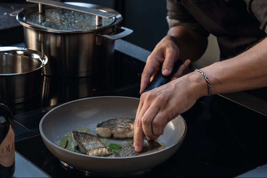 Eine Person benutzt eine Zange, um Fischfilets in einer Fissler-Bratpfanne zu braten; Fissler-Edelstahltöpfe stehen auf den benachbarten Brennern.