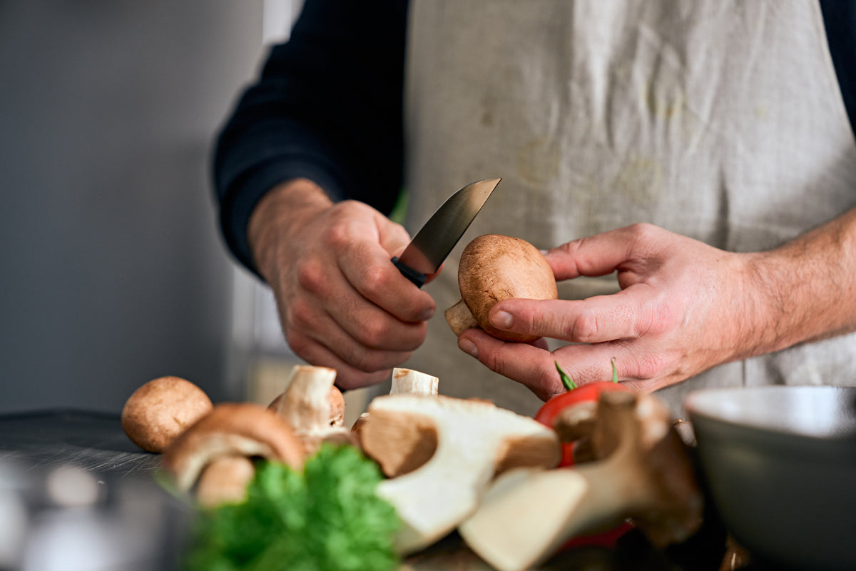 Eine Person, die eine Schürze trägt, schneidet einen braunen Pilz mit dem Fissler ProCut Gemüsemesser (9 cm), das in Deutschland gefertigt wurde und über einen rutschfesten Griff sowie extrem widerstandsfähigen Stahl für präzises Schneiden verfügt. Die Küchenszene zeigt außerdem weitere Pilze, frisches Grünzeug und eine Fissler-Edelstahlschüssel auf der Arbeitsplatte – ein Spiegelbild der seit 1845 bestehenden Tradition von Fissler, innovatives und langlebiges Kochgeschirr herzustellen.