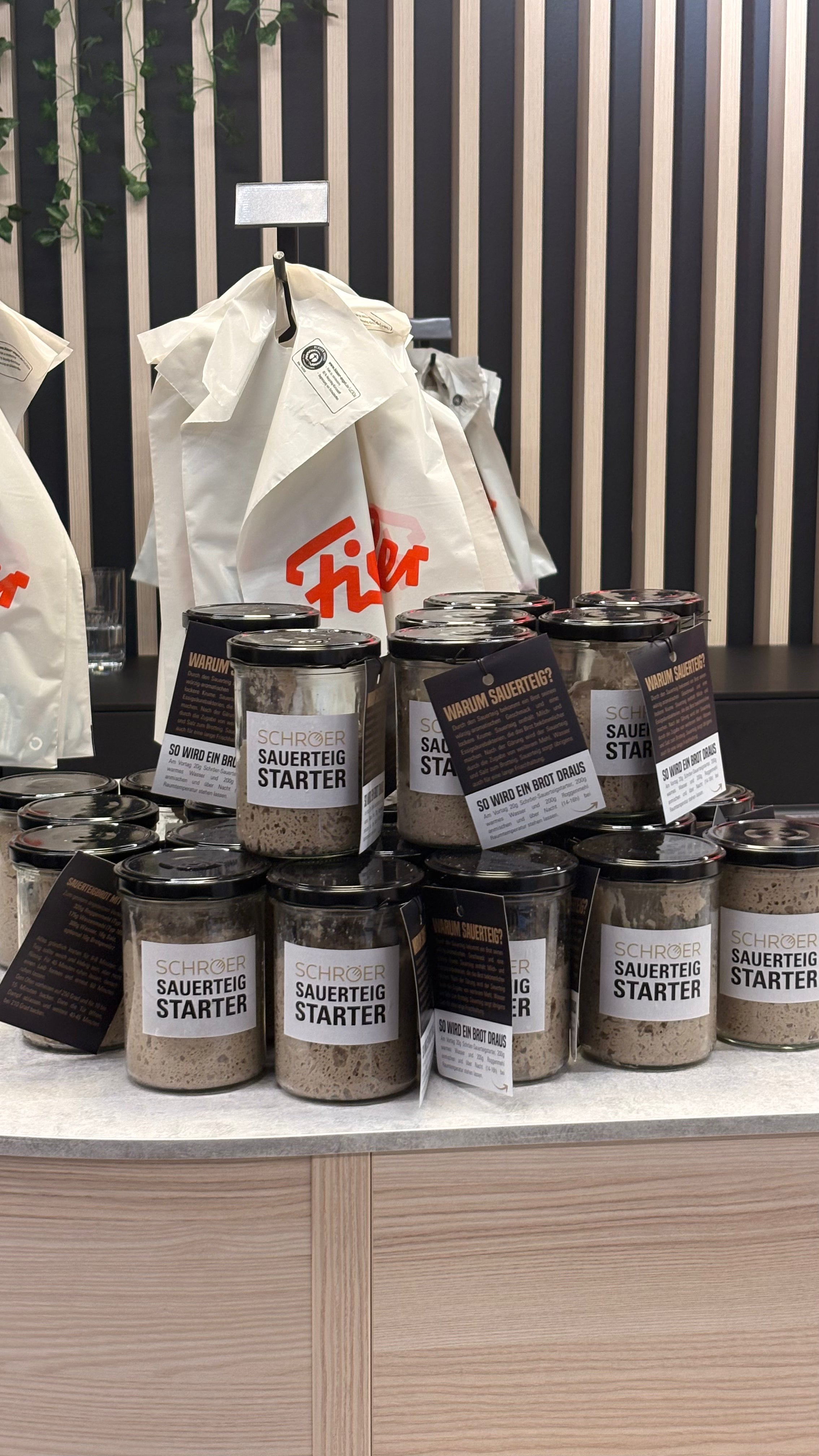 Auf einer Arbeitsplatte stehen ordentlich gestapelte Gläser mit hochwertigem Fissler-Sauerteigstarter, die unsere Tradition von Qualität und Innovation unterstreichen. Im Hintergrund hängen strapazierfähige Tragetaschen mit auffälligen roten Fissler-Logos vor einer eleganten Wand aus schwarzem Holz, die an zeitloses deutsches Design und Handwerkskunst erinnert.