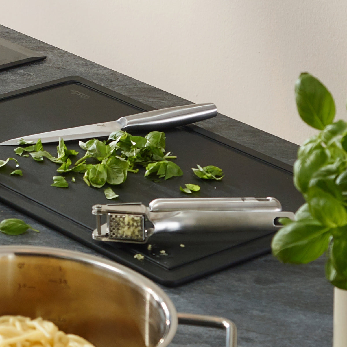 Der Fissler Essential®-Topf mit Nudeln, Knoblauchpresse und frischem Basilikum zeigt auf einer modernen Küchenzeile deutsches Präzisionsdesign.