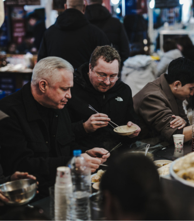 Zwei Männer genießen in einem belebten Markthallenrestaurant eine Mahlzeit mit Essstäbchen, umgeben von einer Vielzahl köstlicher Gerichte, die in stilvollen Schalen und Tassen serviert werden. Die einladende Tischszene spiegelt die Essenz des Zusammenseins und des gemeinsamen Genießens wider und ist inspiriert von Fisslers Engagement für hochwertige kulinarische Erlebnisse. Während der Schwerpunkt auf authentischer Küche liegt, erinnert die Atmosphäre an Fisslers Tradition präzise gefertigter