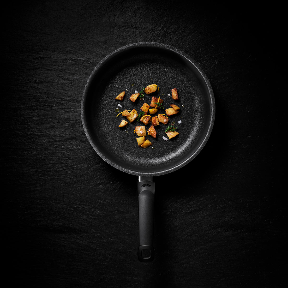 Eine schwarze Bratpfanne von Fissler, die mit präziser Technik für Langlebigkeit und zeitloses Design gefertigt wurde, präsentiert auf ihrer edlen, dunklen, strukturierten Oberfläche einige goldbraun gebratene Kartoffelwürfel, garniert mit frischen Kräutern – ein Beispiel für deutsche Qualität und Innovation bei Kochgeschirr.