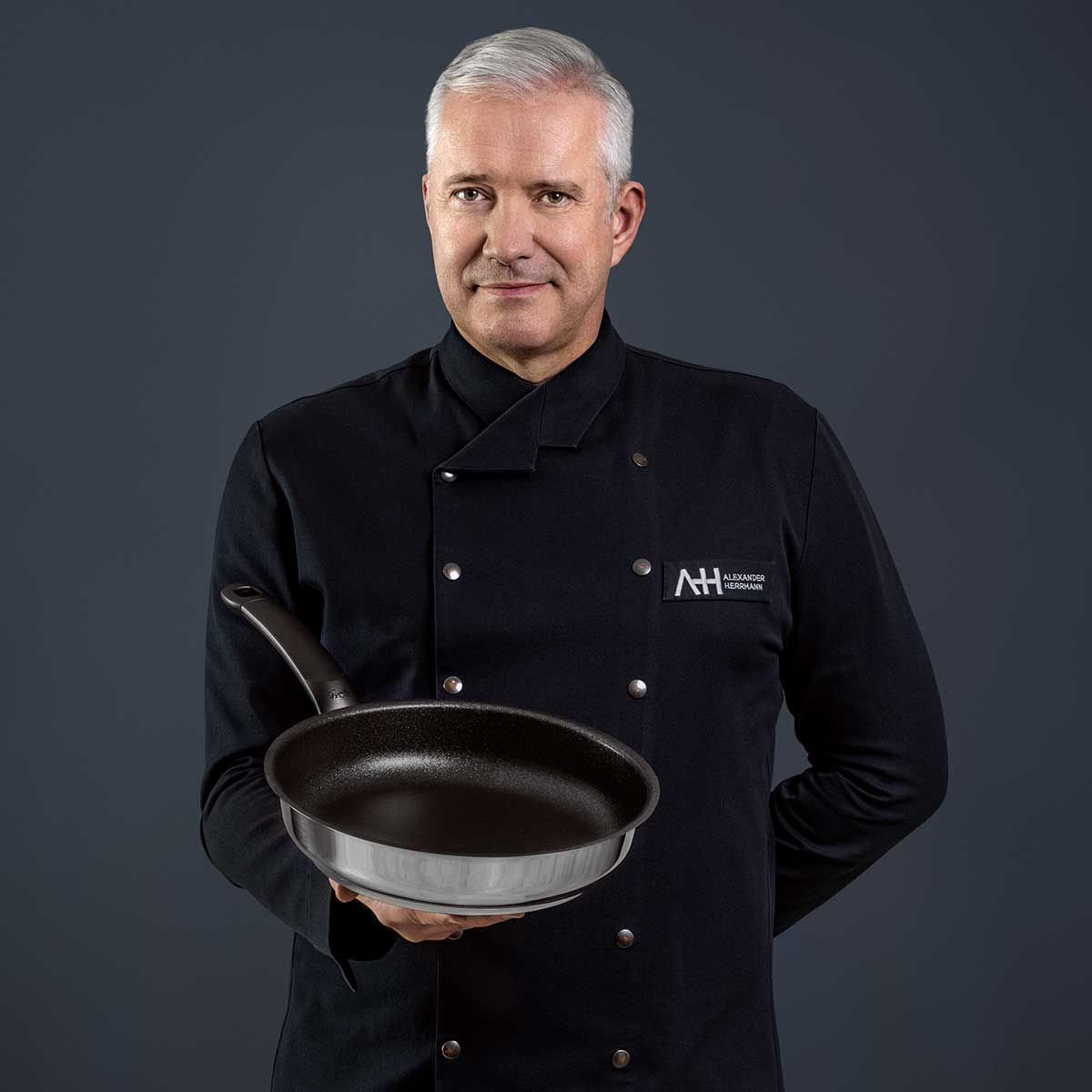 Ein Chefkoch in Schwarz präsentiert die Fissler Adamant® Premium Pfanne - Made in Germany, kratzfest und auf Langlebigkeit getrimmt.
