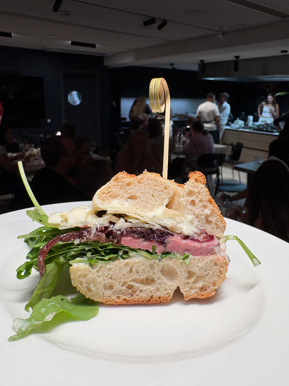 Eine Nahaufnahme eines Gourmet-Sandwichs mit Schichten aus Salat, Käse und Fleisch auf rustikalem Brot, das ordentlich mit einem Holzspieß fixiert ist. Das Sandwich wird elegant auf einem makellos weißen Teller präsentiert, in einer Küchenumgebung, in der hochwertiges Kochgeschirr und Küchenzubehör – Markenzeichen der renommierten deutschen Handwerkskunst von Fissler – dezent im sanft unscharfen Hintergrund zu sehen sind.