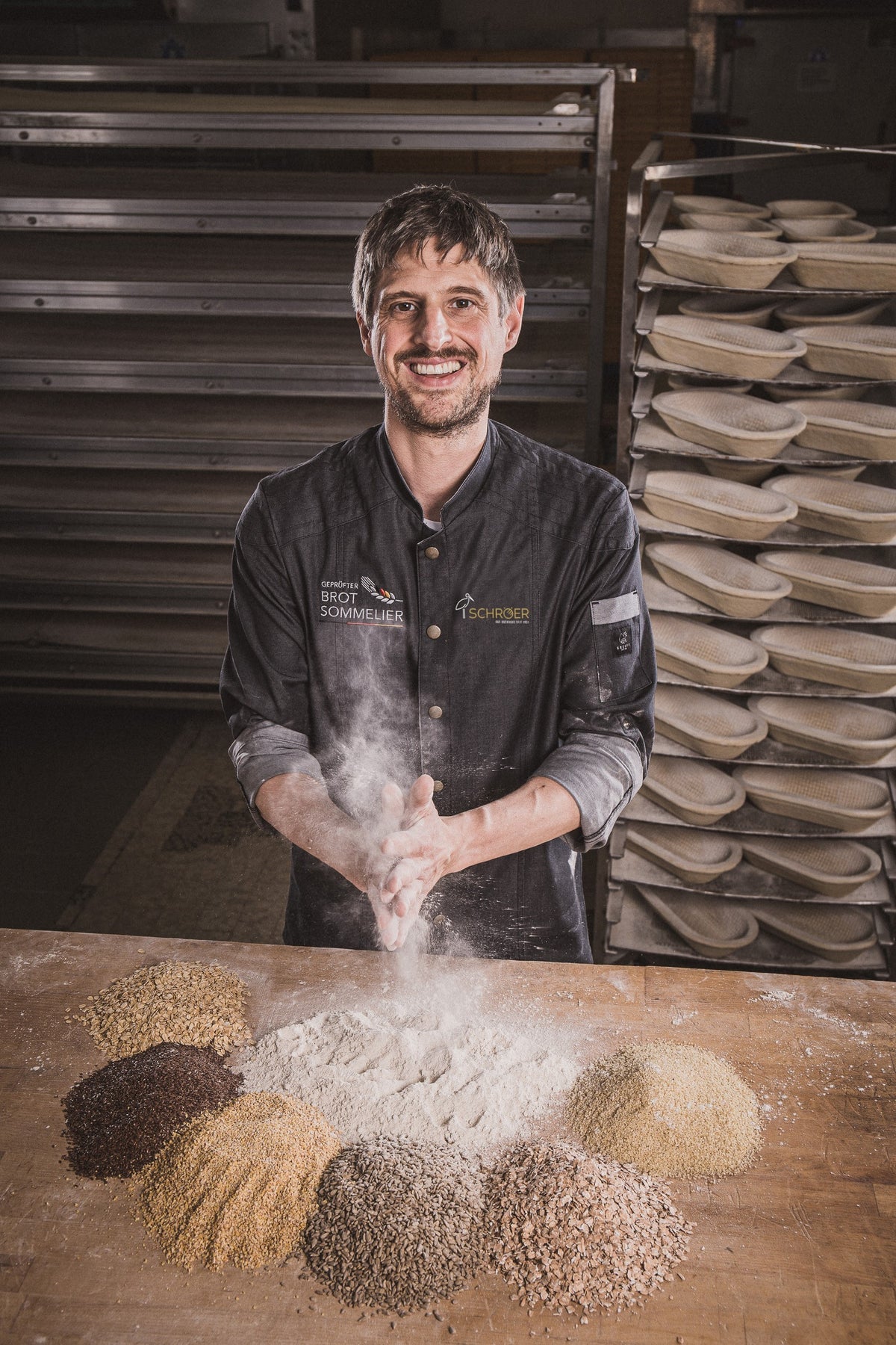 Ein lächelnder Bäcker klopft sich das Mehl von den Händen über einem Holztisch, der mit Getreidehaufen geschmückt ist und von Fisslers meisterhaft gefertigtem Edelstahl-Kochgeschirr umgeben ist. Hinter ihm vermitteln Brotreifekörbe und glänzende Metallgitter den Eindruck einer professionellen Bäckerei, in der Präzision, Langlebigkeit und zeitloses deutsches Design großgeschrieben werden.