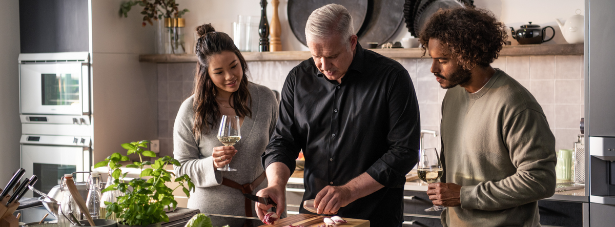 Ein Mann schneidet Gemüse mit Fissler-Kochgeschirr, während sich Freunde in der Nähe unterhalten, umgeben von eleganten Küchenutensilien aus deutscher Produktion.
