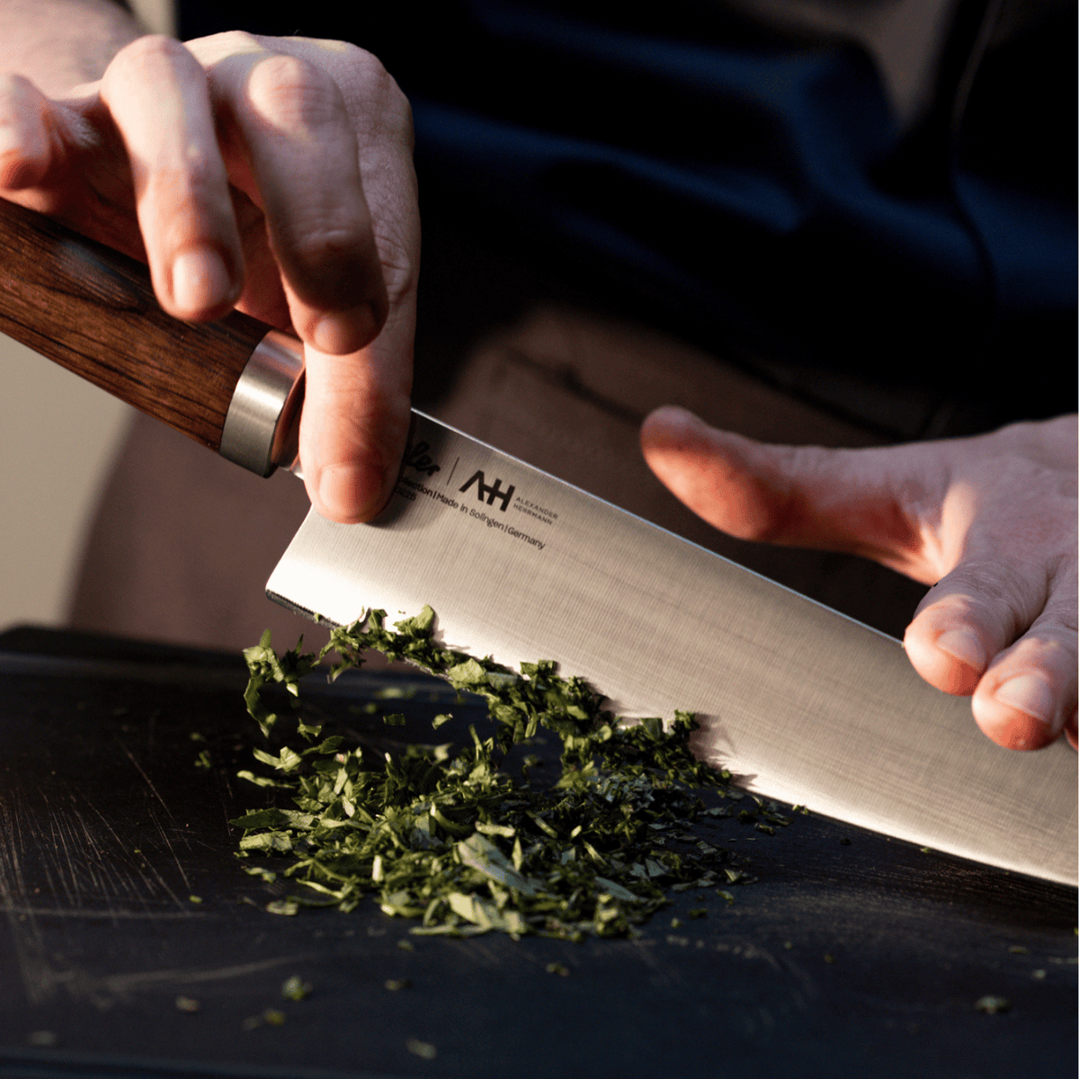 Eine Person hackt fachmännisch frische Kräuter mit einem Präzisionskochmesser von Fissler auf einem schwarzen Schneidebrett, das deutsche Handwerkskunst demonstriert.