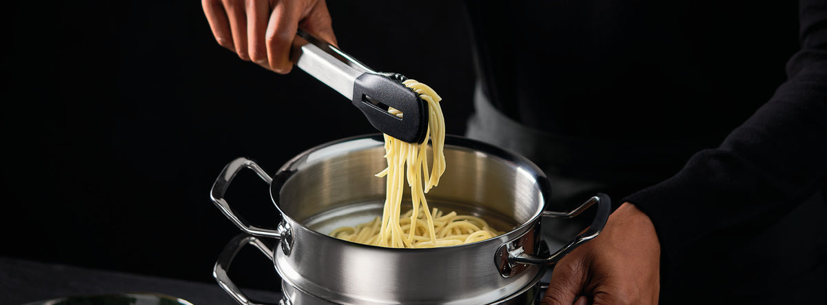 Eine Person hebt Spaghetti mit einer Zange aus einem Fissler-Edelstahltopf und unterstreicht damit dessen deutsche Qualität und zeitloses Design.