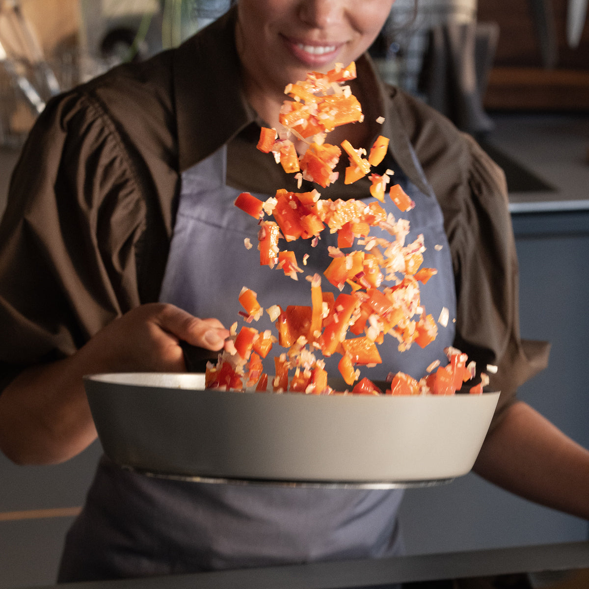 Eine Person in einer Schürze lächelt, während sie Paprika und Zwiebeln in einer hochwertigen Fissler-Edelstahlpfanne in einer modernen Küche brät.