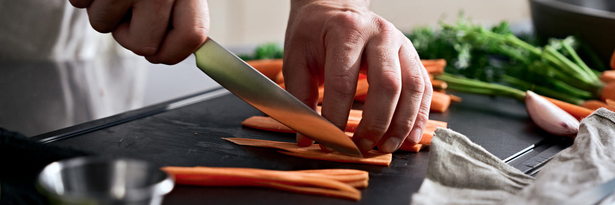 Nahaufnahme von Händen, die mit Präzision Karotten auf einem Schneidebrett schneiden, umgeben von frischem Obst und Gemüse - wie geschaffen für die Fissler-Küche.
