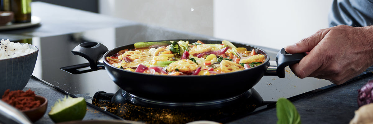 Ein Koch bereitet in einer schwarzen Fissler-Pfanne auf dem Herd ein lebhaftes Rührbraten zu, daneben werden Reis und frische Zutaten präsentiert.