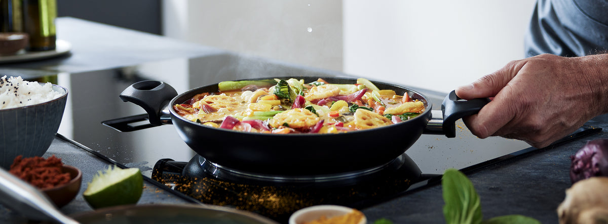 Eine Fissler-Pfanne mit lebhaft gebratenem Gemüse und Garnelen auf dem Herd, umgeben von frischen Zutaten auf einer glatten Theke.