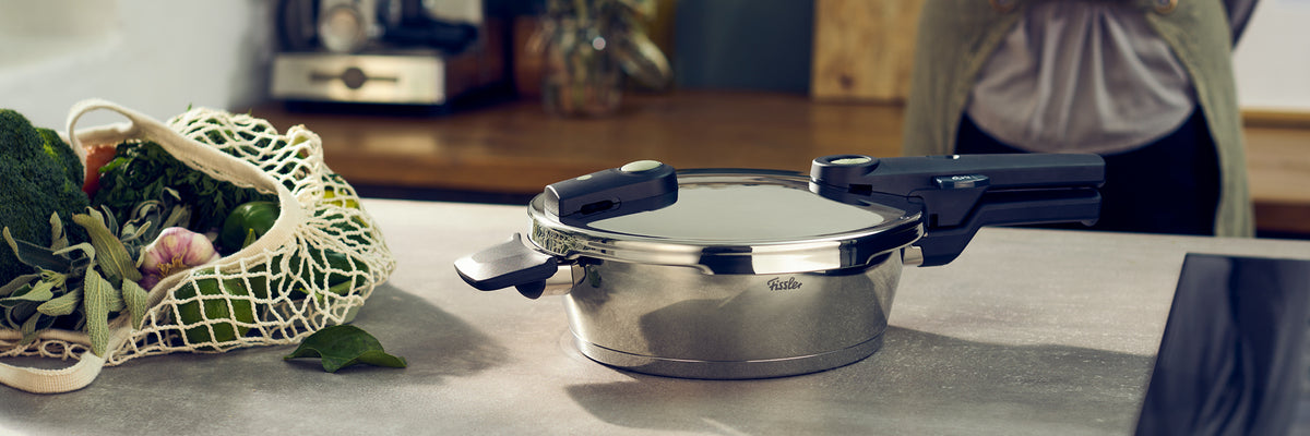 Ein Fissler Schnellkochtopf aus rostfreiem Stahl, Deckel geschlossen, auf einem Tresen neben einem Netzbeutel mit Gemüse; Person unscharf im Hintergrund.