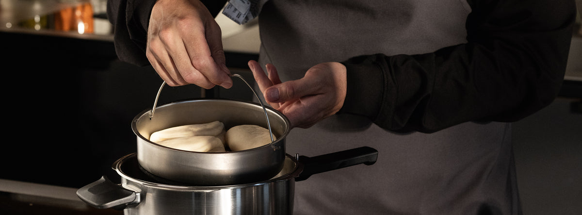 Eine Köchin in grauer Schürze verwendet gestapelte Fissler-Edelstahltöpfe zum Dämpfen von Teig, die Präzision und zeitloses deutsches Design demonstrieren.