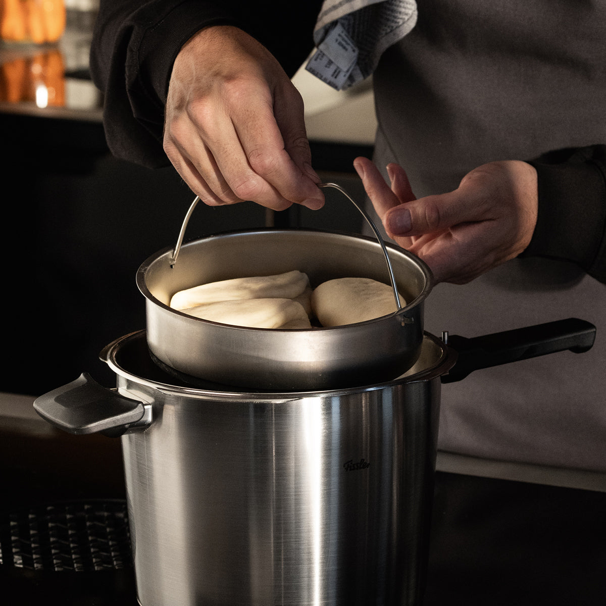 Eine Person hebt einen Dämpfkorb mit Teigbrötchen aus einem Fissler-Edelstahltopf, der deutsche Präzision und zeitloses Design demonstriert.