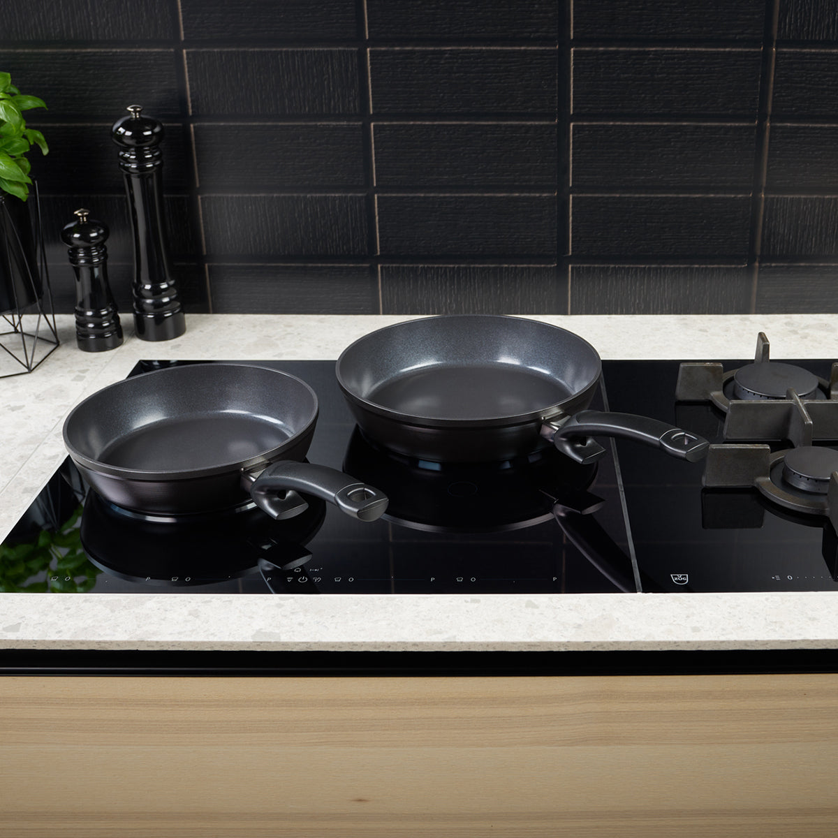 Zwei hochwertige schwarze Fissler-Pfannen, gefertigt mit präziser Technik und zeitlosem deutschem Design, stehen auf einer eleganten Glaskochfläche. Daneben sorgen elegante schwarze Pfeffer- und Salzmühlen für einen Hauch von Raffinesse, ergänzt durch einen Hauch von Grün und eine moderne schwarze Fliesenrückwand im Hintergrund.