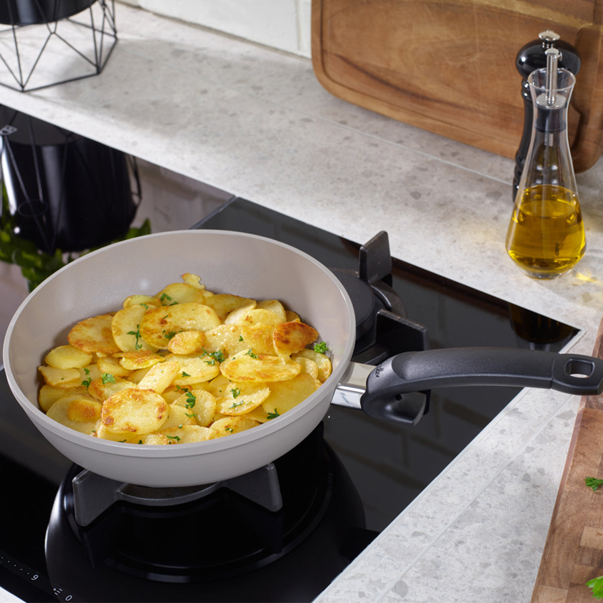 Auf dem Herd steht eine hochwertige Fissler-Pfanne mit präzise gegarten, goldbraunen Kartoffelscheiben, die das Bekenntnis der Marke zu deutscher Ingenieurskunst und zeitlosem Design unterstreicht. Daneben ergänzen eine Glasflasche mit Olivenöl, eine Pfeffermühle und ein Holzschneidebrett die elegante Funktionalität des Küchenarbeitsbereichs auf einer hellen Arbeitsplatte.