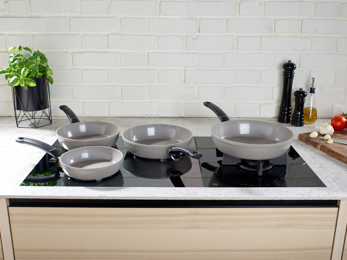 Vier Fissler Ceratal® Comfort Pfannen auf einer eleganten Herdplatte, mit frischen Kräutern, Gewürzen und Produkten neben ikonischem deutschen Design.
