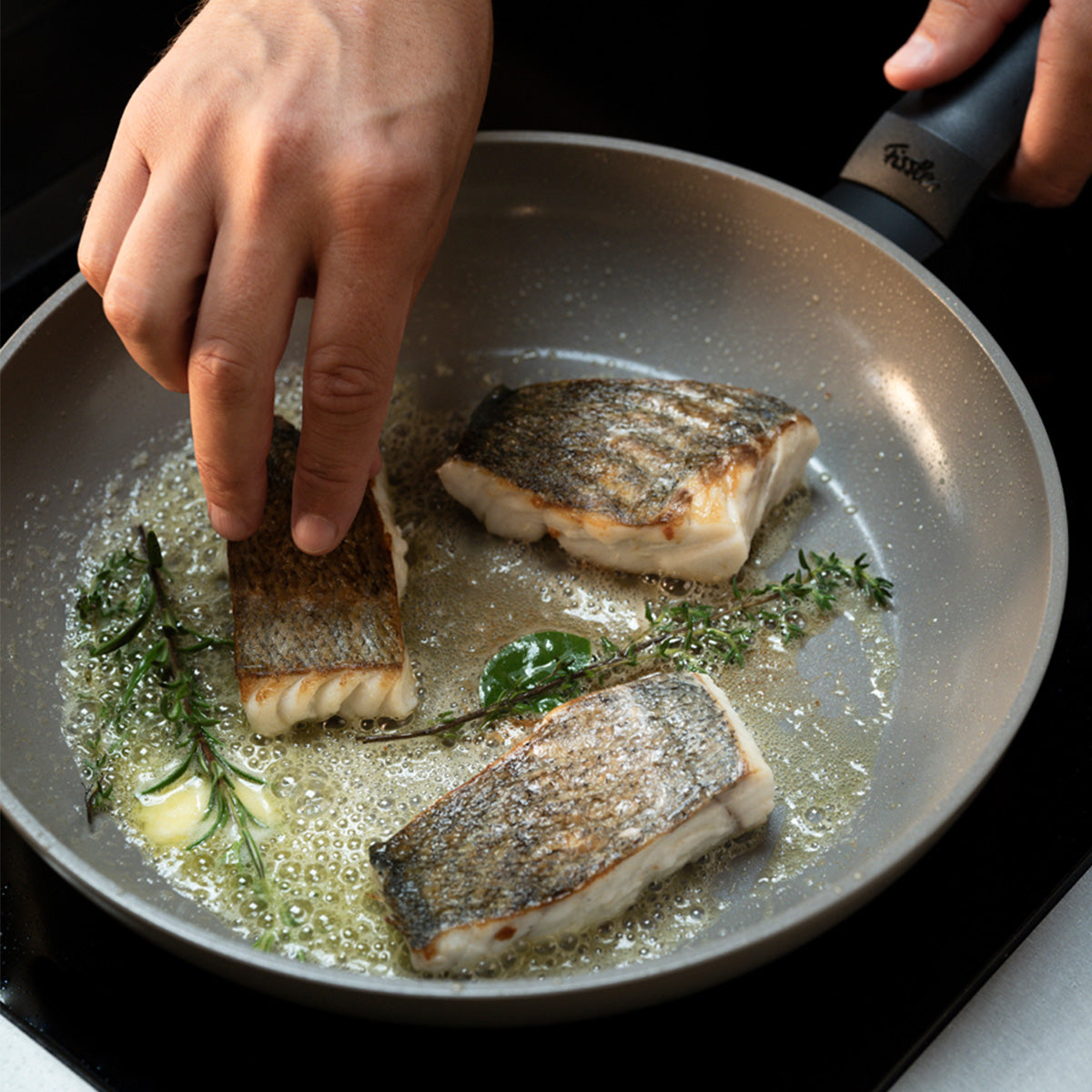 Erleben Sie Präzisionskochen mit der Fissler Ceratal® Evo Classic Pfanne: Drei Fischstücke mit Haut werden fachmännisch mit Kräutern und Butter zubereitet, während eine Hand ein Filet sorgfältig zurechtstutzt. Hervorragende deutsche Ingenieurskunst und Innovation sorgen für eine gleichmäßige Wärmeverteilung und damit für perfekte Ergebnisse – ein Markenzeichen der langen Tradition von Fissler im Bereich hochwertiger Kochgeschirre seit 1845.
