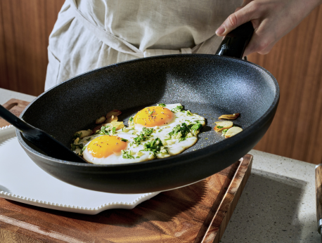 Eine Person in einer Schürze hält eine hochwertige Fissler-Pfanne mit zwei Spiegeleiern, frisch gehackten Kräutern und Knoblauch in der Hand, bereit, das Gericht auf einen Teller zu servieren. Die Szene verdeutlicht deutsche Präzisionstechnik und zeitloses Design im kulinarischen Alltag.