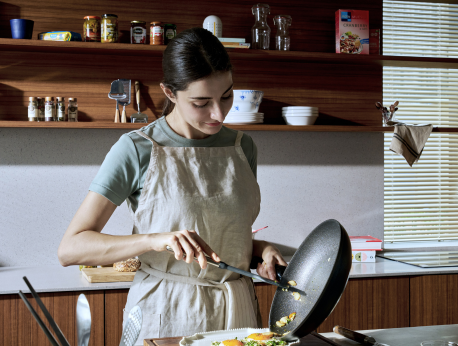 Eine Frau in einer Schürze serviert mit einem Pfannenwender Speisen aus einer hochwertigen Fissler-Edelstahlpfanne auf einen Teller in einer modernen Küche mit Holzregalen und einer Auswahl an frischen Zutaten – ein Bild, das die präzise Technik und das zeitlose Design der Fissler-Kochgeschirre widerspiegelt.