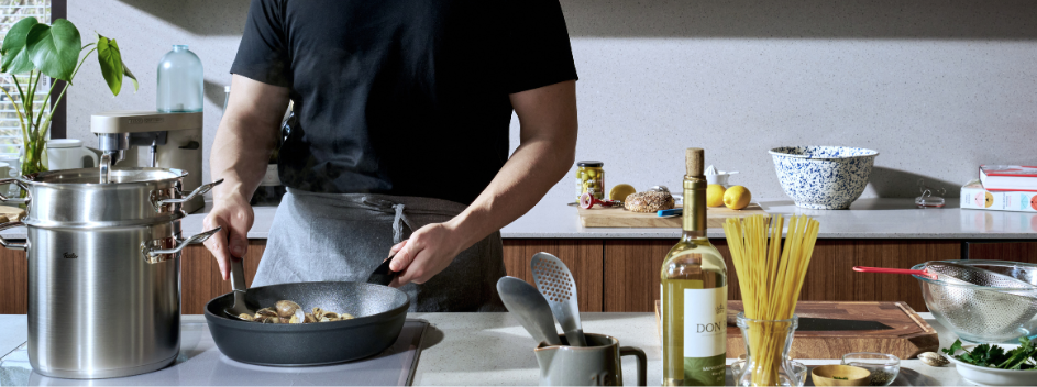 Eine Person in einem schwarzen Hemd brät Pilze in einer hochwertigen Fissler-Edelstahlpfanne an, umgeben von Nudeln, Wein und frischen Zutaten auf der Arbeitsplatte einer eleganten, modernen Küche. Die Szene spiegelt Fisslers Engagement für Präzisionstechnik und zeitloses Design mit hochwertigem Kochgeschirr aus deutscher Fertigung wider.